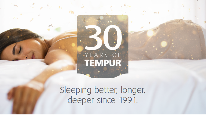 Vrouw die op haar buik ligt en het bed waar ze op ligt omhelst. Boven haar valt gouden confetti naar beneden en staat het logo van 30 jaar tempur, sleeping better, longer and deeping since 1991.